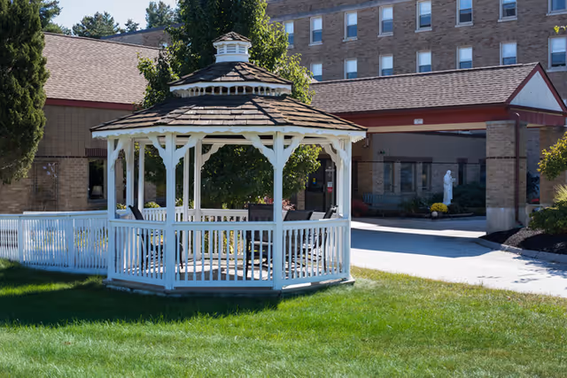 White wooden gazebo with a shingled roof situated on a green lawn in front of a multi-story brick building with a covered entrance. Trees and shrubs surround the area, and a white fence is visible on the left side.
