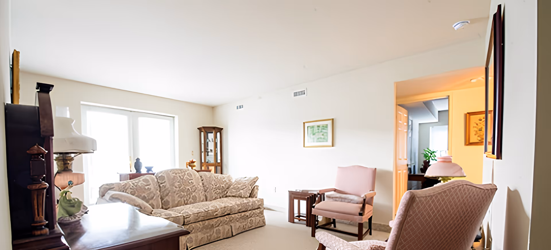 A bright living room with a patterned beige sofa, two upholstered armchairs, a wooden side table, a grandfather clock, and a glass display cabinet. The room has white walls, a large window letting in natural light, and framed artwork on the walls.