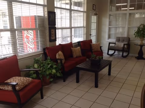 A bright sitting area with red cushioned chairs and a matching sofa arranged around a black coffee table. There are several potted plants, large windows with white blinds, and a Coca-Cola vending machine visible outside the window. The floor is tiled and there is a rocking chair and a small round table with a plant in the background.