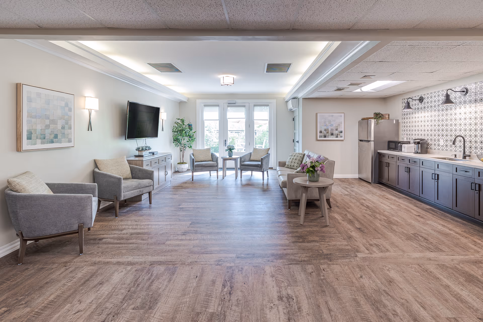 A bright and spacious common area in a senior living facility featuring a seating area with gray armchairs and a sofa, a wall-mounted TV, and a kitchenette with a refrigerator, microwave, coffee maker, and sink. The room has wood flooring, light-colored walls, and large windows with glass doors leading outside.