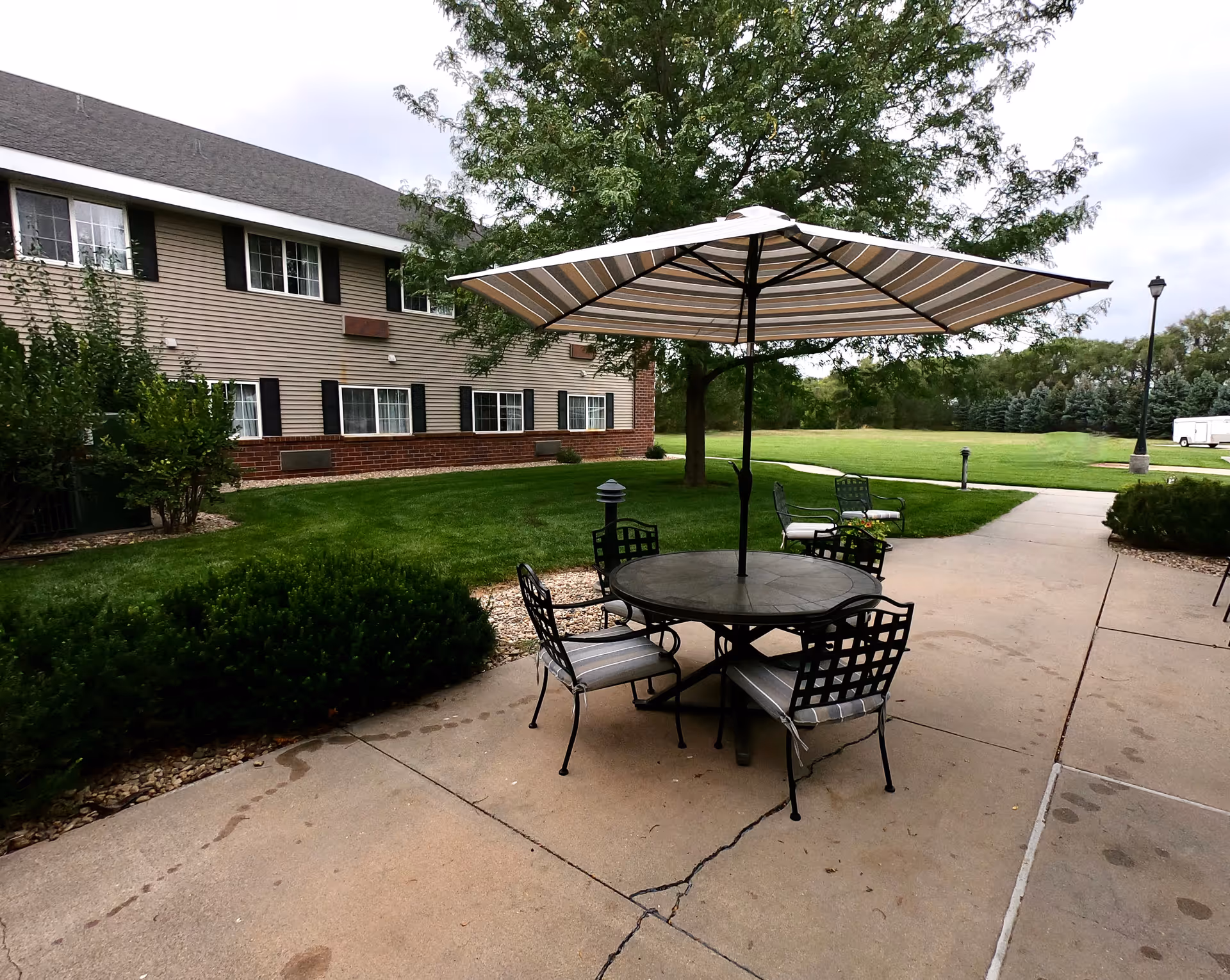 Outdoor patio area at Grandview Assisted and Independent Living featuring a round table with four metal chairs with cushions and a large striped umbrella. The patio is surrounded by green grass, bushes, and trees, with a two-story building with multiple windows in the background.