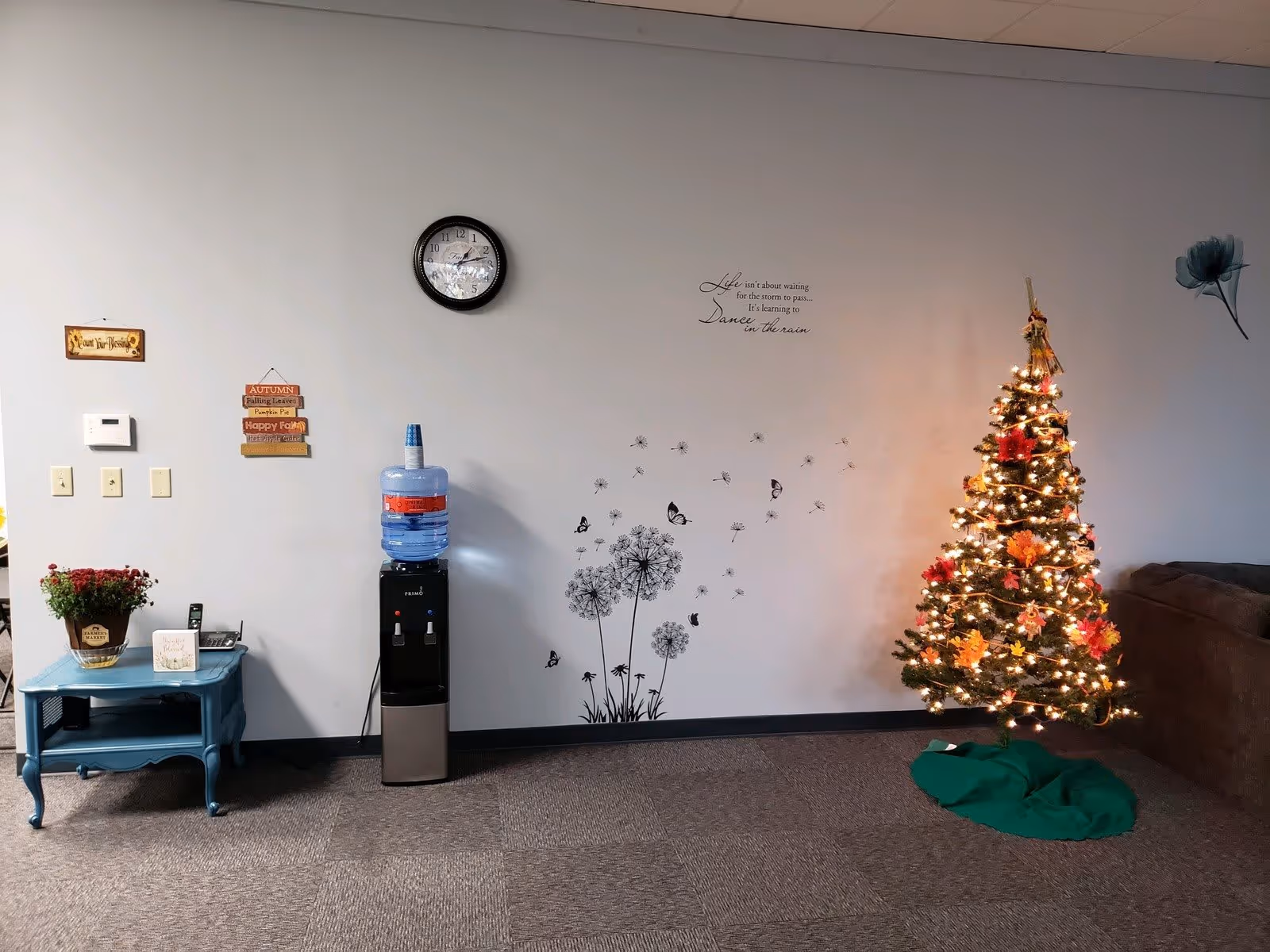 Interior of a room with a decorated Christmas tree with lights and autumn leaves on the right, a brown couch partially visible, a wall clock, a water dispenser with a large water bottle, a small blue table with a potted plant and a phone, and wall decals including dandelions, butterflies, a flower, and an inspirational quote.
