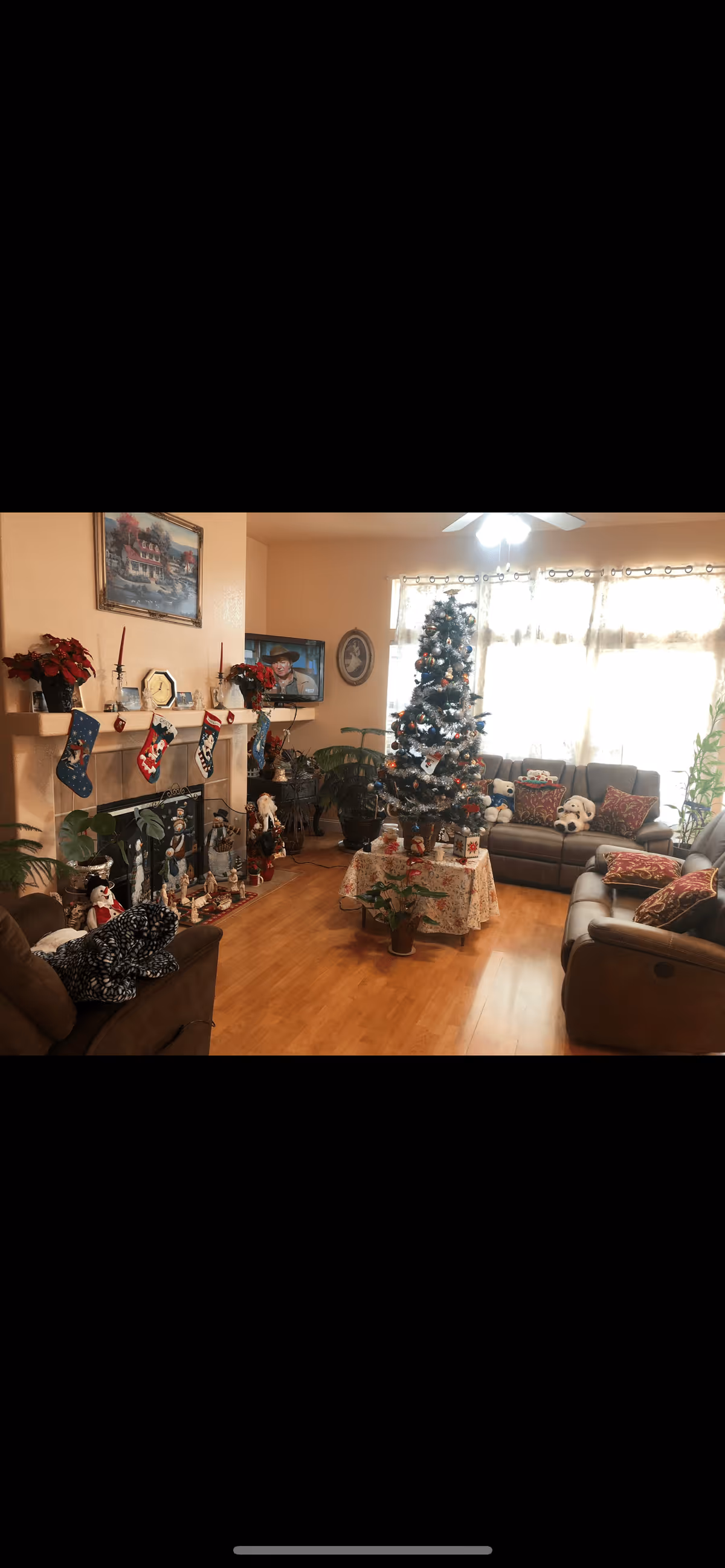 A cozy living room decorated for Christmas with a decorated Christmas tree in the center on a table covered with a cloth. The room has brown leather sofas with patterned cushions, a fireplace with Christmas stockings hanging on the mantle, and various holiday decorations including poinsettias and figurines. Large windows with sheer curtains allow natural light to fill the room.
