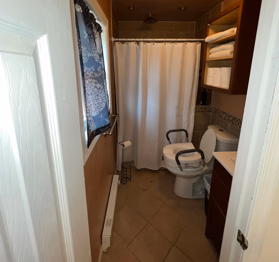 A small bathroom with a white door partially open, a window with a patterned curtain on the left wall, a toilet with arm supports, a white shower curtain, a tiled floor, and a wooden cabinet with shelves holding towels and toilet paper.