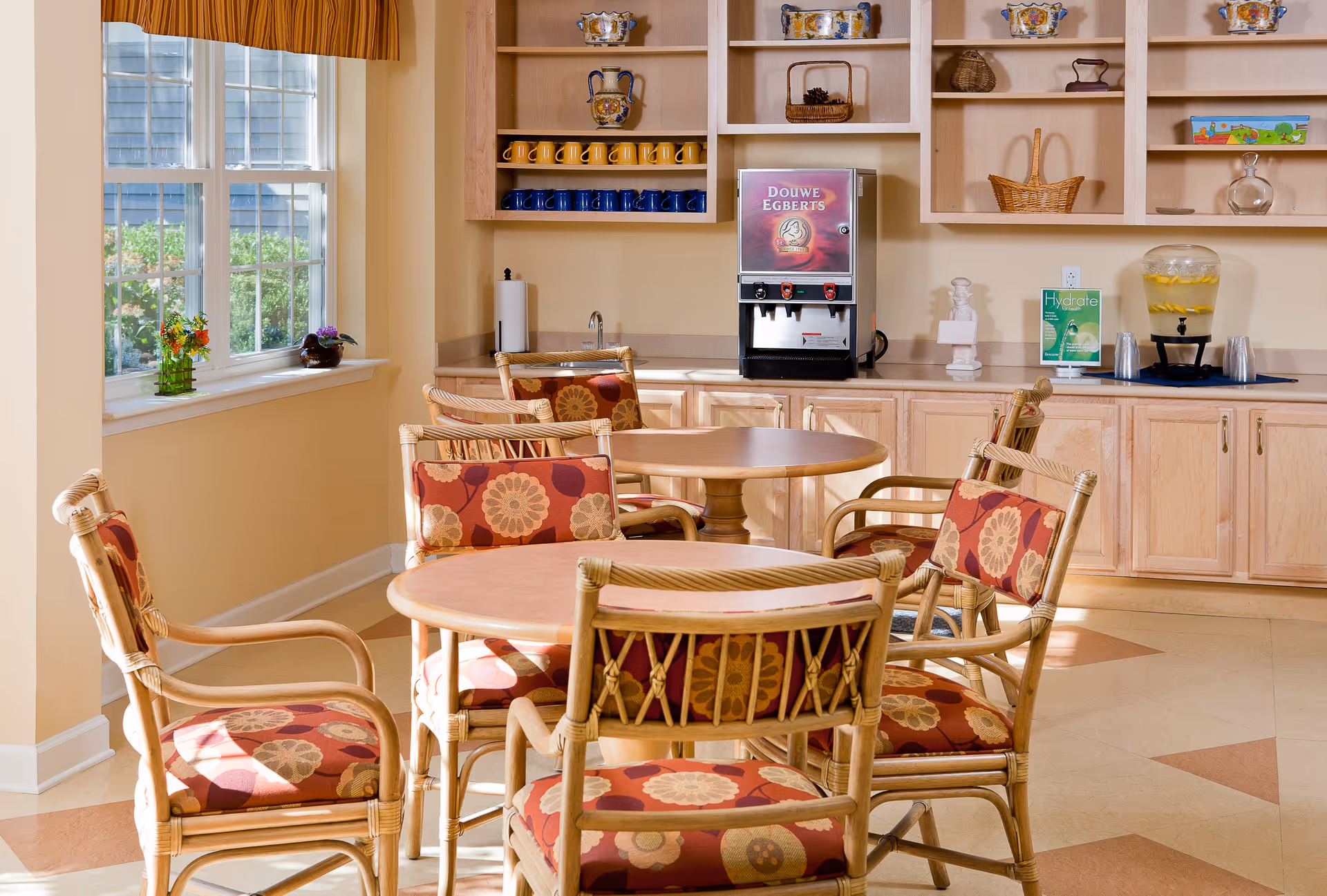 A bright room with round wooden tables and wicker chairs with floral patterned cushions. There is a large window with a yellow valance letting in natural light. On the back wall, there are wooden cabinets and shelves holding decorative items, yellow and blue mugs, a Douwe Egberts coffee machine, and a water dispenser with lemon slices. A small sign about hydration is also visible on the counter.