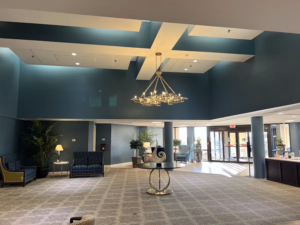 Spacious senior living community lobby with high ceilings, a large modern chandelier, patterned carpet, blue walls, seating areas with chairs and sofas, potted plants, and large glass entrance doors letting in natural light.