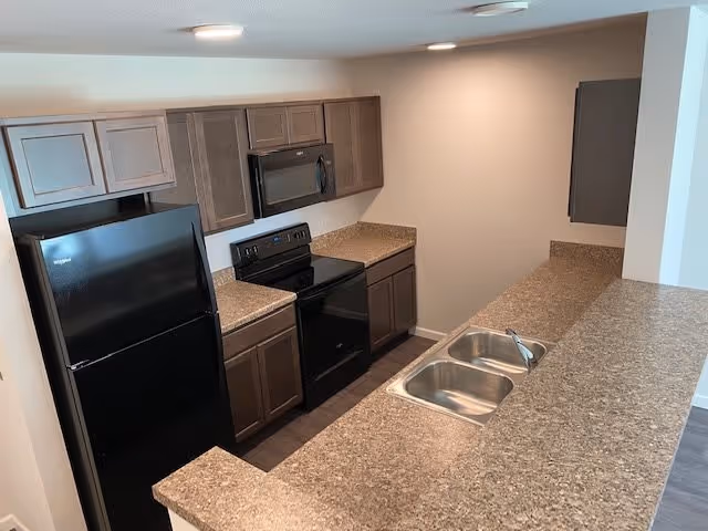 A modern kitchen with brown cabinets, a black refrigerator, black stove with oven, black microwave, and a double stainless steel sink set in a brown speckled countertop. The kitchen has a neutral color scheme with beige walls and dark wood flooring.
