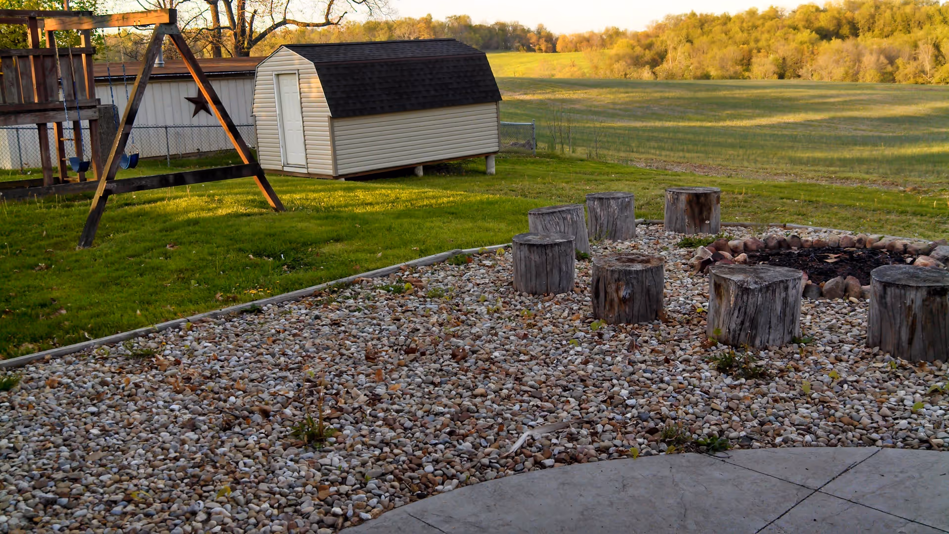 Outdoor area with a small shed, a wooden swing set, a fire pit surrounded by tree stump seats, and a grassy field with trees in the background under a clear sky.