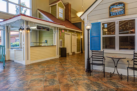 Indoor common area designed to look like a small village street with a yellow building labeled 'Carry Town' and a gray building with blue shutters and a sign reading 'River Road Tavern'. There are two black chairs and a small round table in front of the tavern, a streetlamp, and large windows letting in natural light.