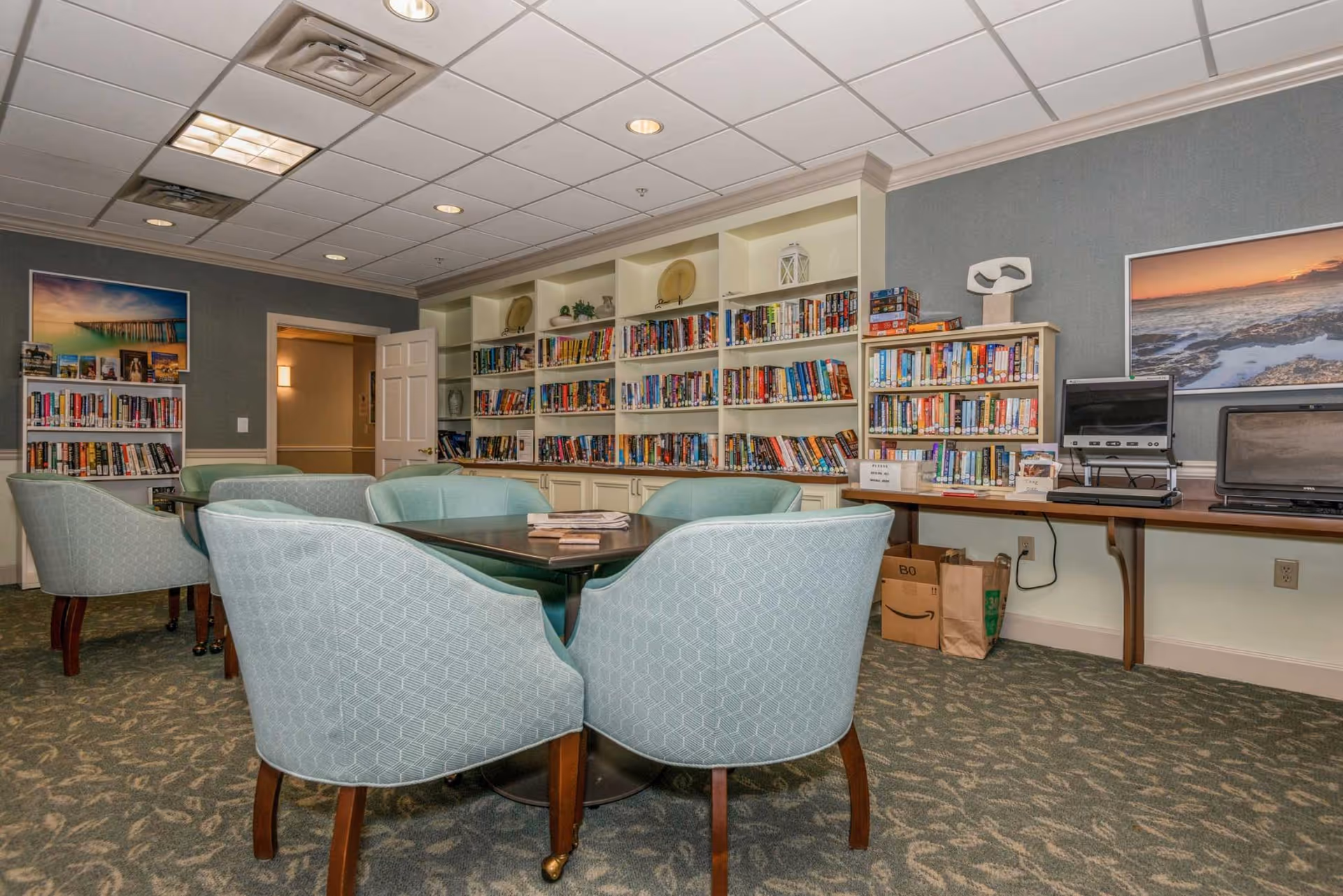 A cozy interior community library with bookshelves, a table surrounded by upholstered chairs, and computers on a side desk.