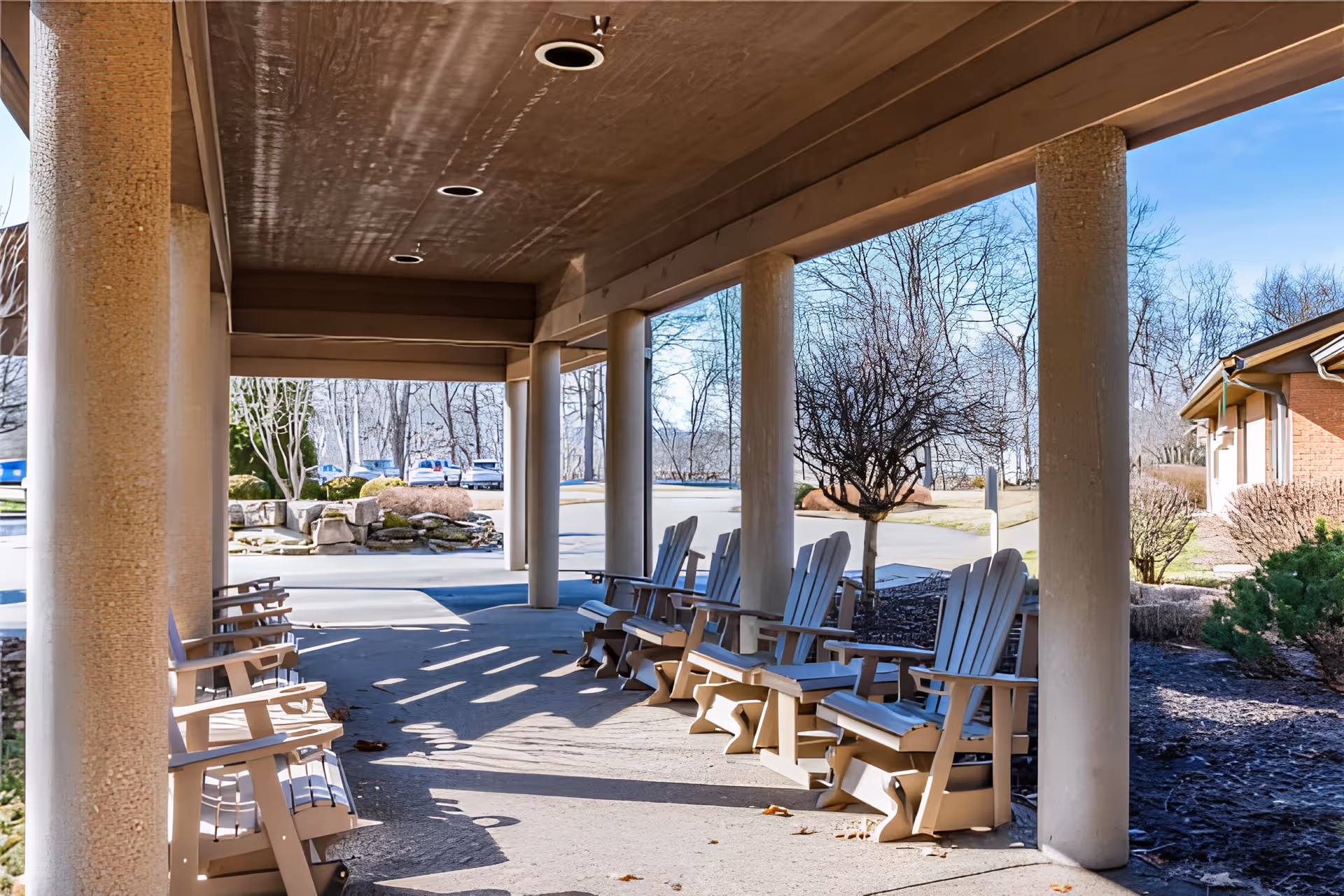 Covered outdoor patio with rows of wooden Adirondack chairs under columns facing a driveway and landscaped grounds.