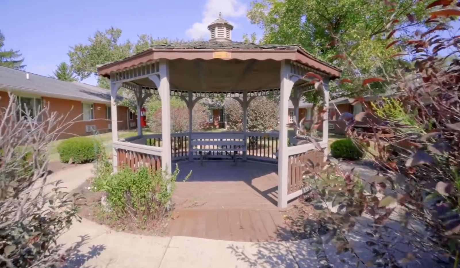 A wooden gazebo with a bench inside, surrounded by bushes and trees, located in an outdoor garden area of a residential facility with buildings visible in the background under a clear blue sky.