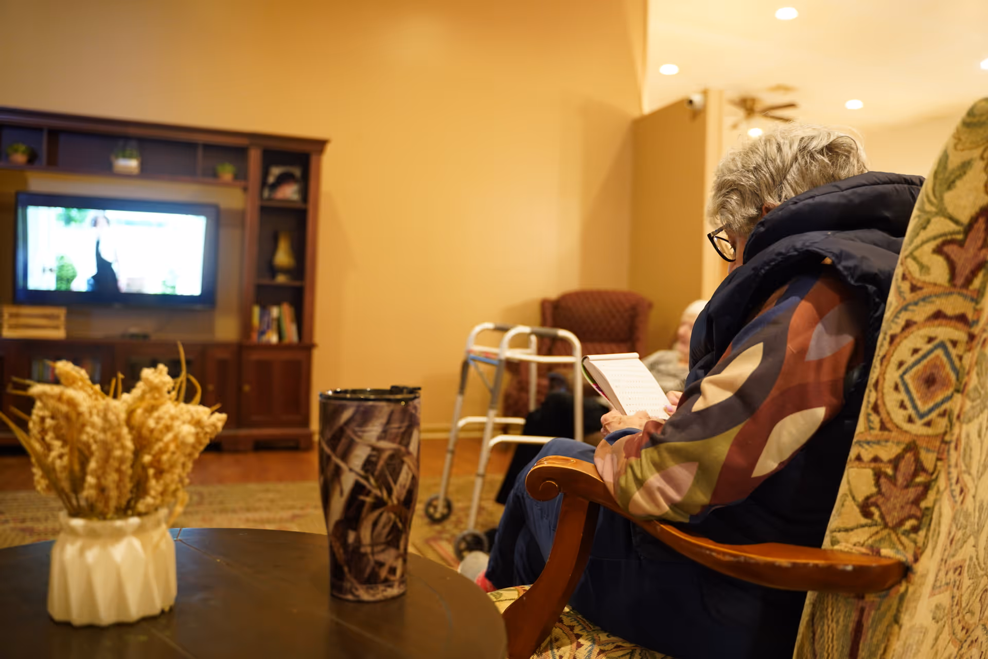 An elderly person sitting in a patterned armchair reading a book in a cozy living room. In the background, there is a wooden entertainment center with a TV, a walker, and another person sitting in a chair. A table with a vase of dried flowers and a travel mug is in the foreground.