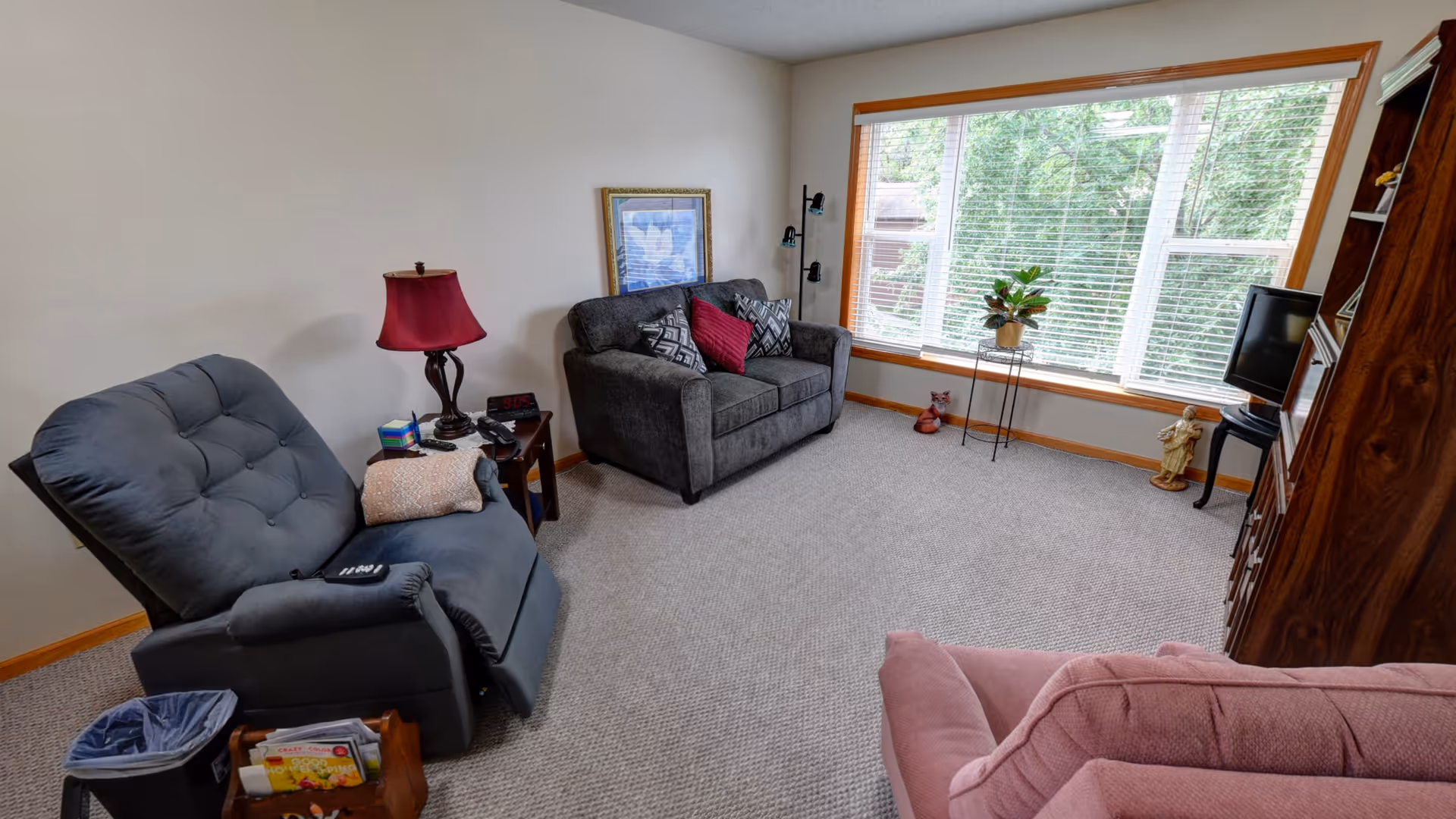 A cozy living room with a large window letting in natural light. The room features a gray loveseat with patterned and solid red pillows, a dark gray recliner chair with a remote on the armrest, a pink armchair, a wooden side table with a red lamp, a digital clock, and a phone. There is a wooden cabinet with a small TV on a stand, a plant on a metal stand near the window, and decorative figurines on the floor. The carpet is light beige, and the walls are plain white.