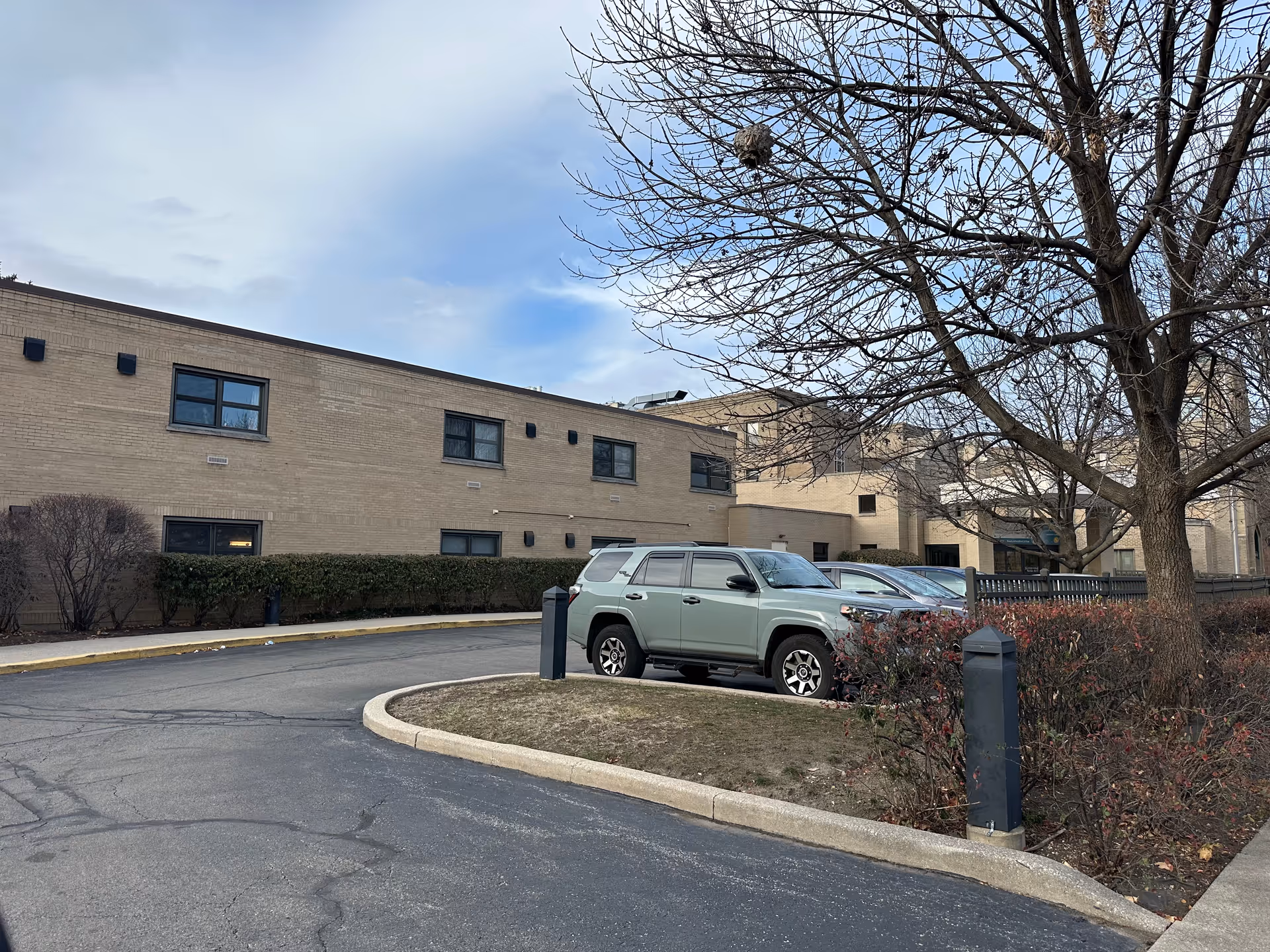 Exterior view of a beige brick building with multiple windows, a parking area with a few parked cars, leafless trees, and some shrubs under a partly cloudy sky.