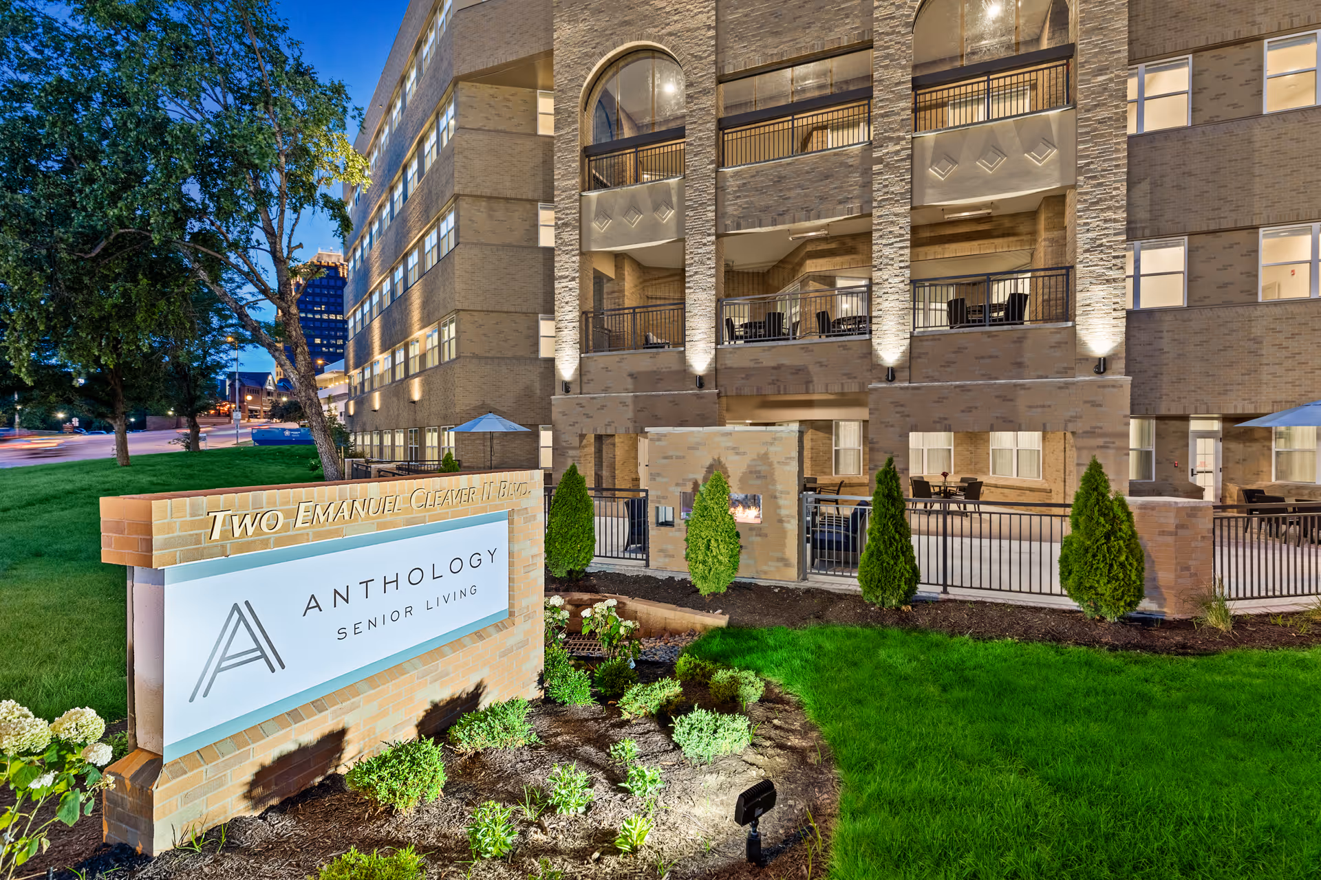 Exterior view of a senior living facility building at dusk with well-lit balconies and a landscaped garden area in front. A large brick sign in the foreground reads 'Two Emanuel Cleaver II Blvd' and 'Anthology Senior Living'.