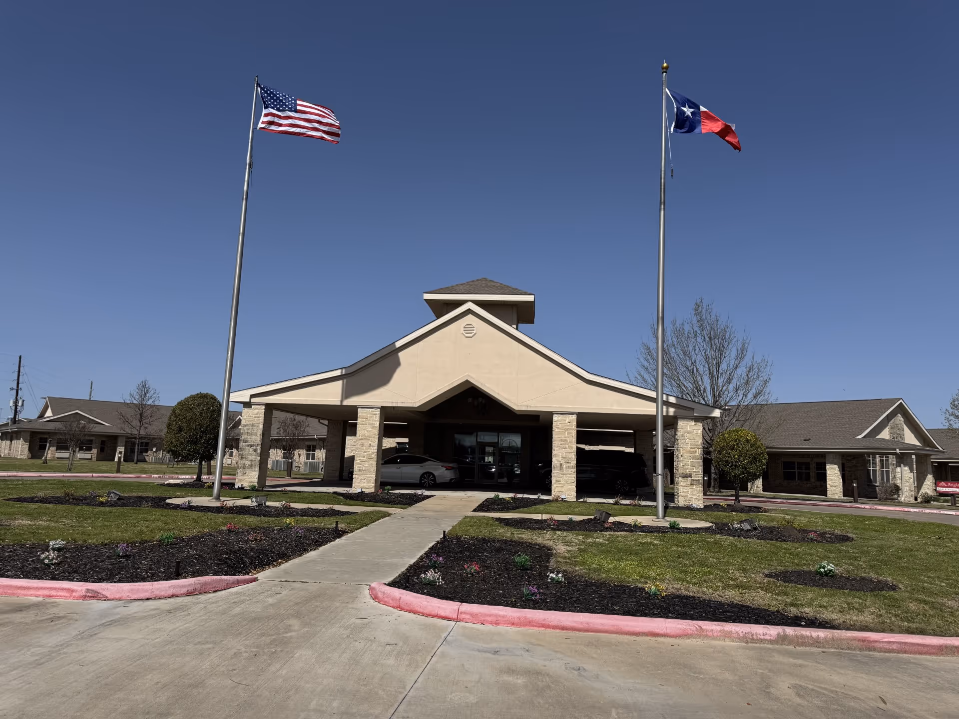 Front exterior view of Eagle Crest Rapid Recovery facility with two flagpoles displaying the American flag and the Texas state flag, a covered entrance with stone pillars, and a clear blue sky.