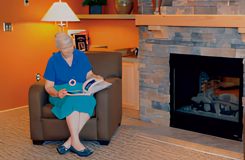 An elderly woman sitting in a brown armchair reading a book in a cozy living room with an orange wall, a lit table lamp, built-in shelves with decorative items, and a stone fireplace.