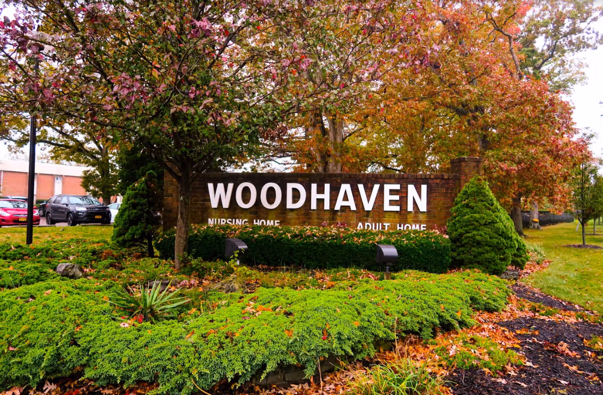 Brick entrance sign reading "WOODHAVEN" surrounded by landscaped shrubs and autumn trees at the facility entrance.