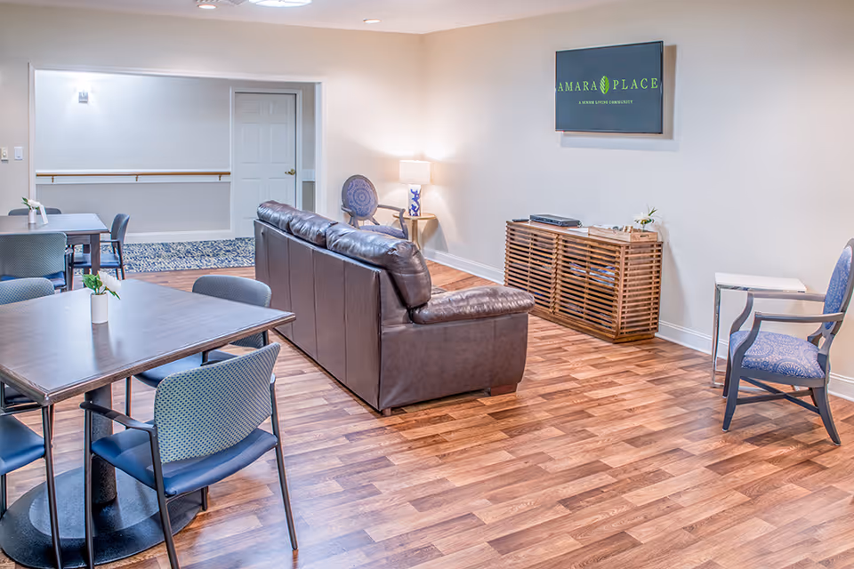A cozy lobby area featuring a brown leather couch, wooden tables, and decorative plants. A television is mounted on the wall, and there are chairs arranged around the space.