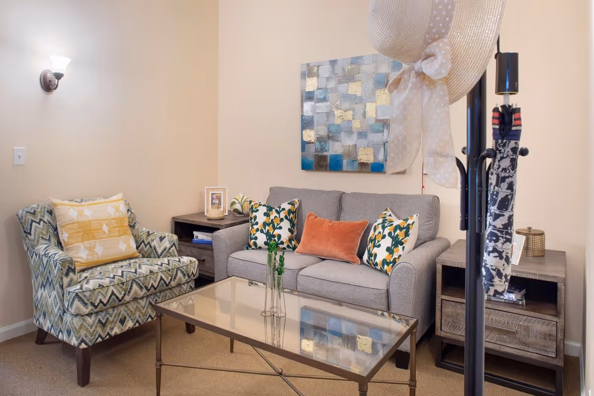 A cozy living room area with a patterned armchair with a yellow and white pillow, a gray sofa with three decorative pillows, a glass coffee table with a small plant, two wooden side tables with decorative items, and a wall-mounted abstract painting. A coat rack with a hat and umbrella is visible in the foreground.
