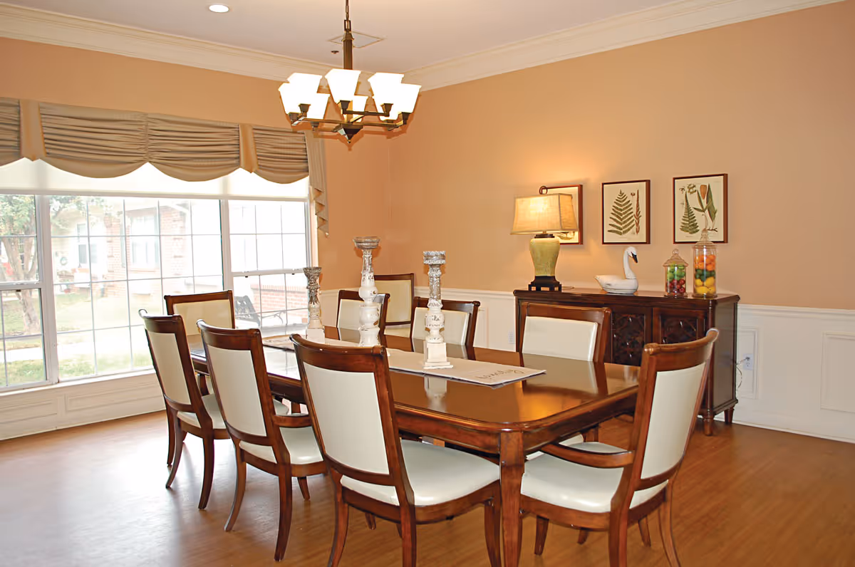 Well-lit dining room with a wooden table and eight chairs, chandelier, sideboard with lamp and decor, and a large window.