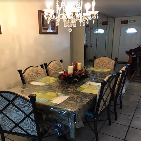 A dining room with a rectangular table covered with a patterned tablecloth and a clear plastic cover. The table is set with placemats, napkins, and glasses. A centerpiece with candles and red decorations is in the middle of the table. There are eight chairs around the table with patterned cushions. A chandelier with lit bulbs hangs above the table. A large mirror covers the wall behind the table, reflecting the room and two white doors with windows.