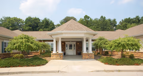 Front exterior view of a single-story building with a covered entrance supported by white columns, surrounded by landscaped greenery and small trees under a partly cloudy sky.