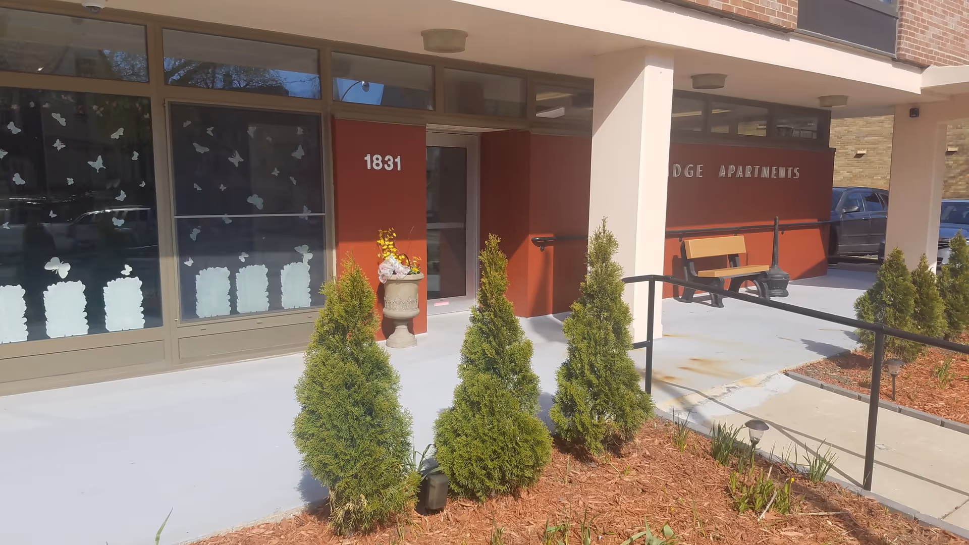 Entrance to Cambridge Apartments building with a red wall displaying the number 1831 and the words 'Cambridge Apartments'. There are small green shrubs planted in mulch in front of the entrance, a bench near the wall, and windows decorated with white butterfly and flower decals.