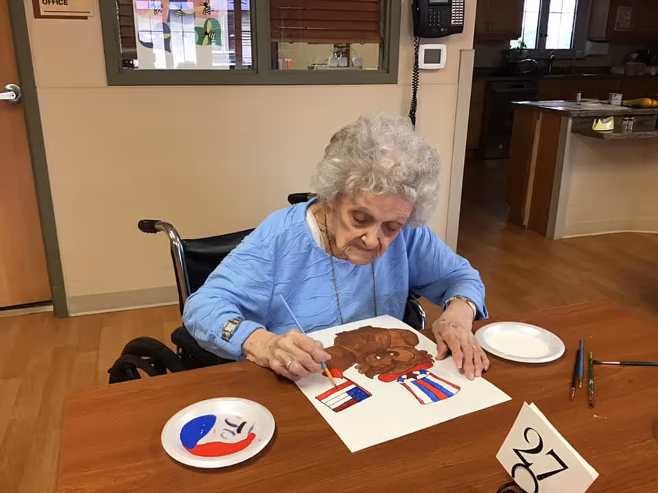 An elderly woman in a wheelchair is painting a picture of a brown dog wearing a red, white, and blue outfit at a wooden table inside a room. There are paintbrushes and paper plates with paint on the table, and a small table number card with the number 27 is visible. The room has wooden floors and a kitchen area in the background.