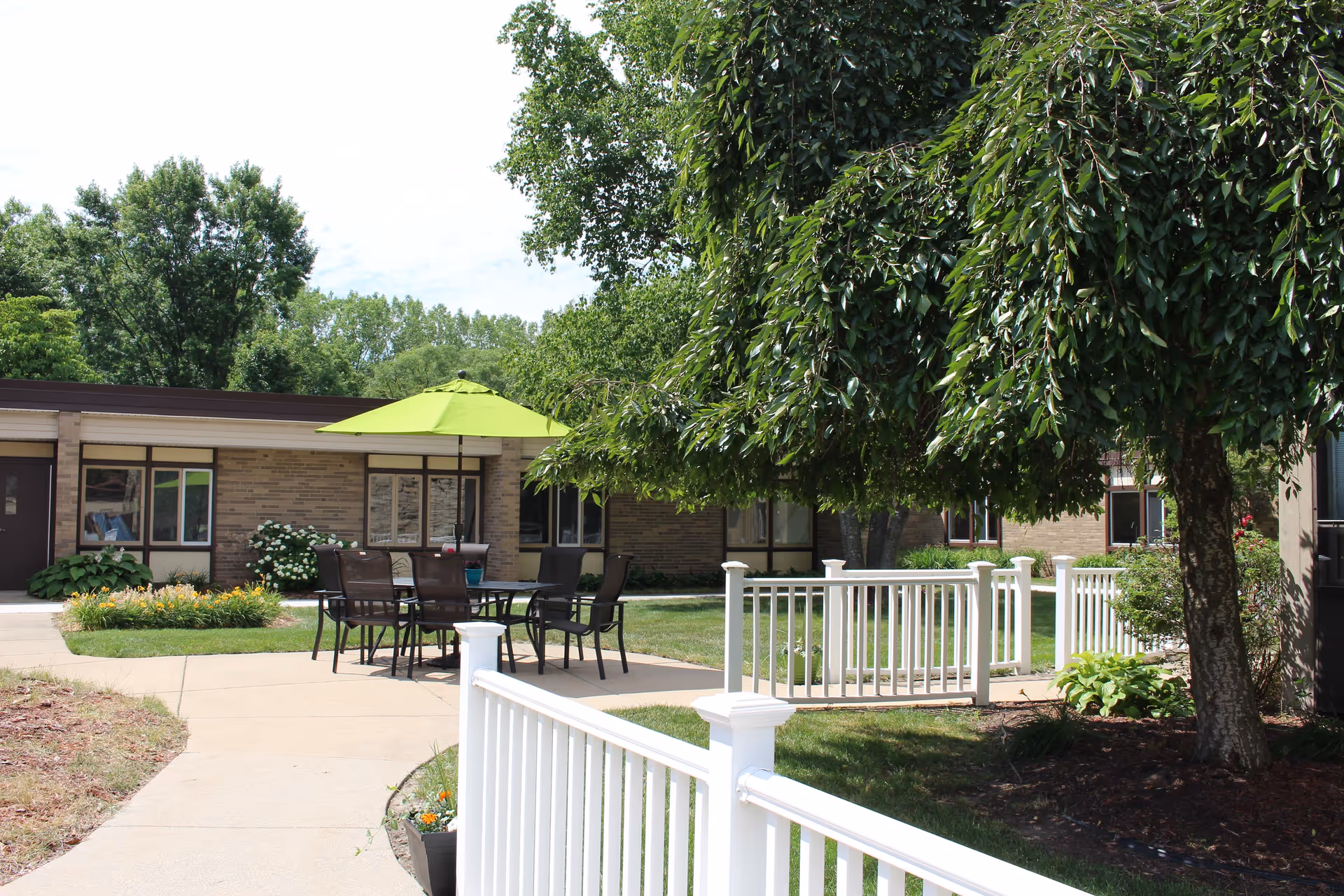 Outdoor courtyard area at MediLodge of Grand Rapids featuring a paved walkway, white railing, a large tree, and a patio table with chairs under a green umbrella. The background shows a single-story brick building with windows and some greenery.