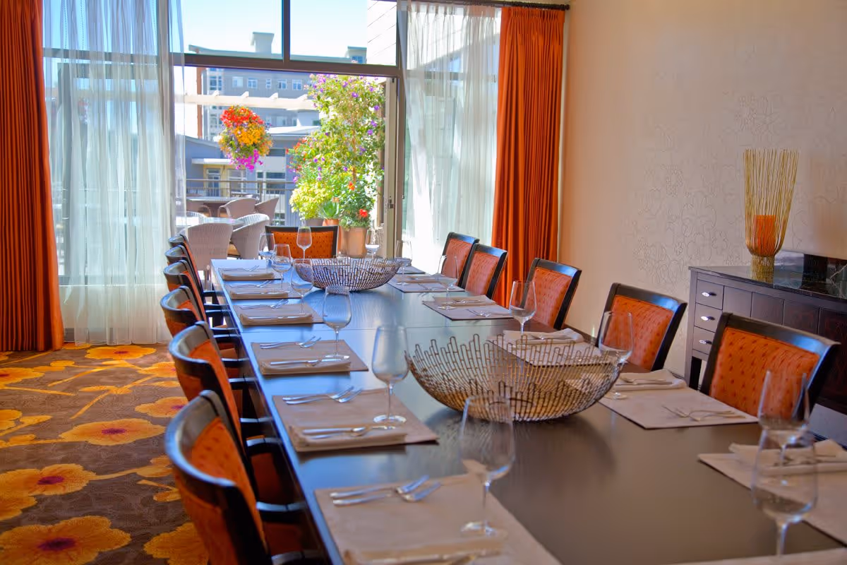 A long dining table set with placemats, napkins, cutlery, and empty wine glasses in a bright room with large windows and orange curtains. Outside the windows, there is a patio area with chairs and colorful hanging flower baskets.