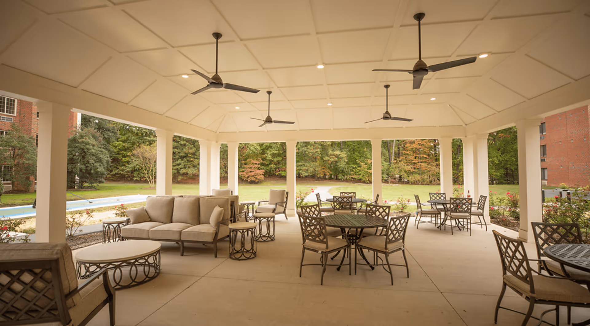 Covered outdoor patio with cushioned seating, multiple dining tables and ceiling fans overlooking landscaped grounds.