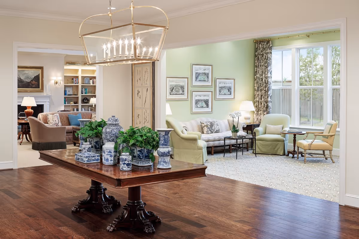 A bright and elegant interior space featuring a wooden table with blue and white ceramic vases and green plants in the foreground. Beyond the table, there is a sitting area with a striped sofa, two armchairs, a side table with a lamp, and framed artwork on a light green wall. Large windows allow natural light to fill the room, and another cozy seating area with a fireplace and bookshelves is visible in the background.