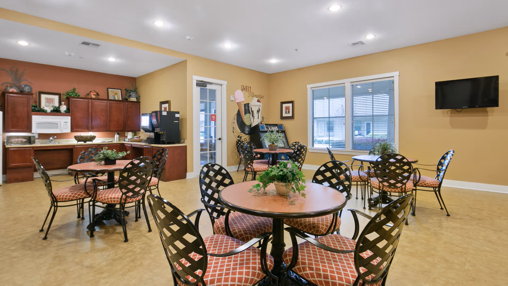 A dining area with several round tables and metal chairs with red patterned cushions. The room has beige walls and a light brown floor. There is a small kitchen area with wooden cabinets, a microwave, and a beverage dispenser. A wall decoration with an ice cream cone and the words 'Sweet Memories' is visible near a glass door. A flat-screen TV is mounted on the wall next to a window with blinds.