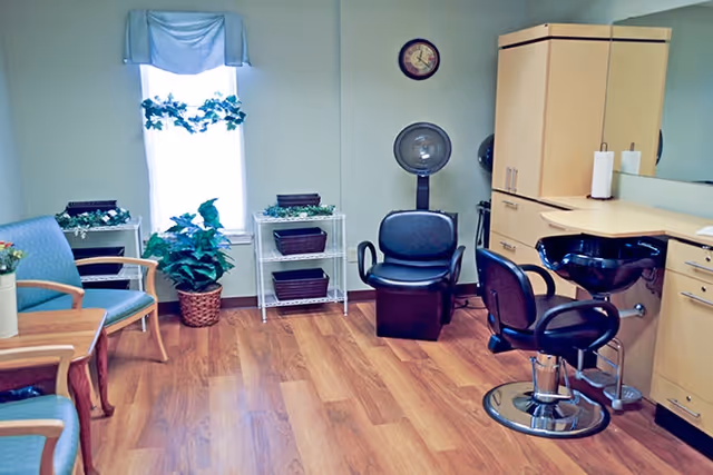 A bright salon room with wooden flooring, featuring a black salon chair and hair washing station on the right, two green cushioned chairs with wooden arms on the left, a small wooden table, a window with a blue valance and green plants on a white shelf and in a basket, and a wall clock above the salon chair.