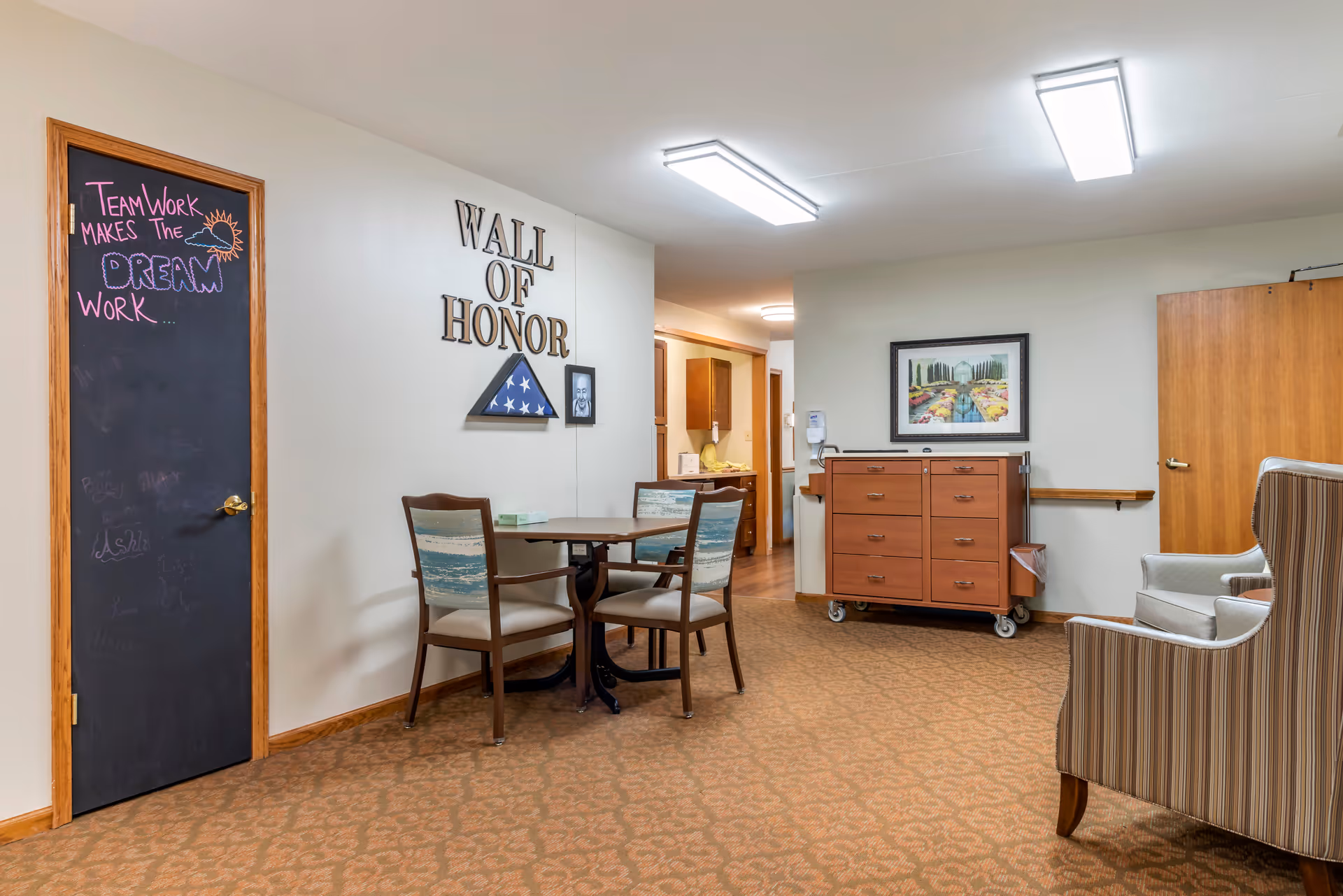 A cozy interior space in a senior living facility featuring a small round table with three chairs, a striped armchair, and a wooden cabinet on wheels. On the wall, there is a 'Wall of Honor' display with a folded American flag and a framed photo. A blackboard door with the phrase 'Team Work Makes The Dream Work' written in colorful chalk is also visible. The room has warm lighting and carpeted flooring.
