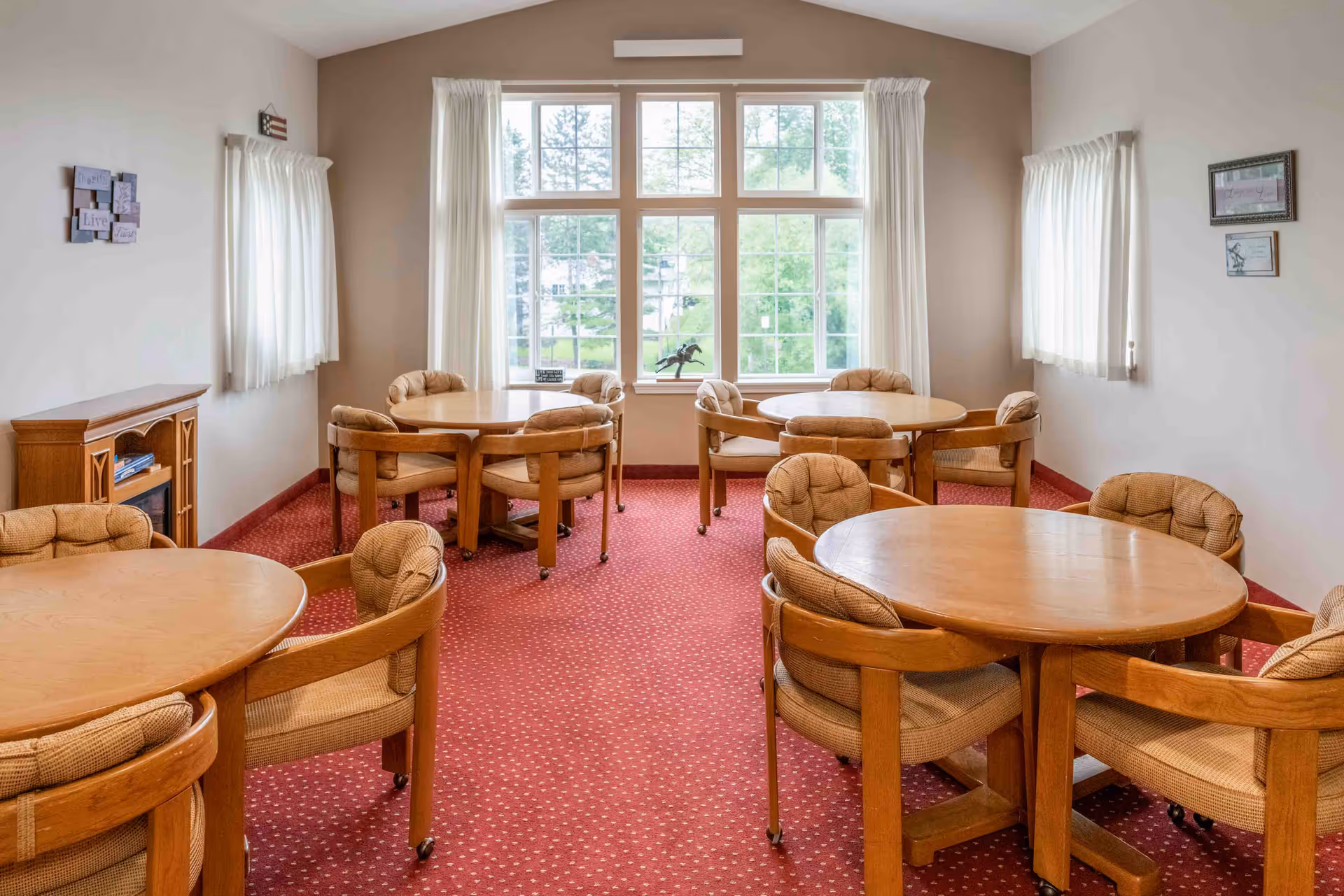 Bright carpeted common room with round wooden tables and cushioned chairs arranged near large windows.