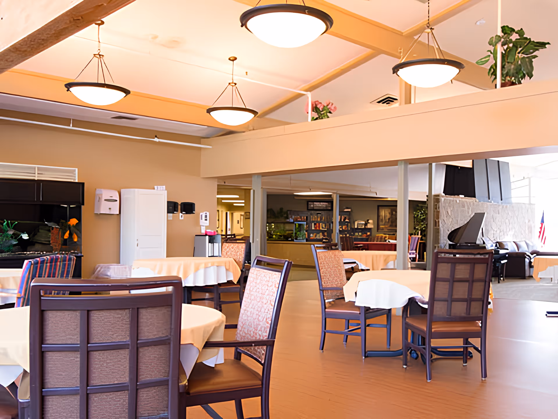 A spacious dining area with round tables covered in beige tablecloths and wooden chairs with cushioned seats. The room has warm lighting from ceiling fixtures and a light brown floor. In the background, there is a stone fireplace, a piano, and some plants, creating a cozy atmosphere.