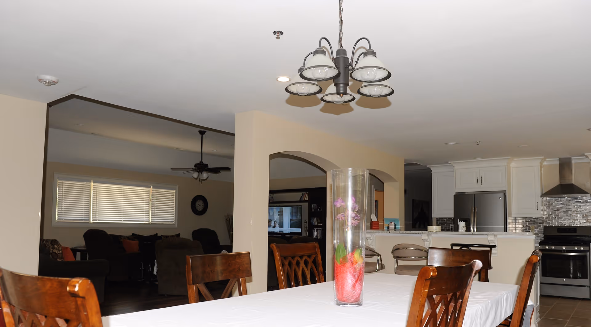 Interior view of a senior living facility showing a dining area with a white tablecloth-covered table and wooden chairs. A tall glass vase with pink flowers and red decorative elements is placed on the table. In the background, there is a kitchen with white cabinets, stainless steel appliances, and a tiled backsplash. Beyond the kitchen, a living room area with dark furniture and a ceiling fan is visible.