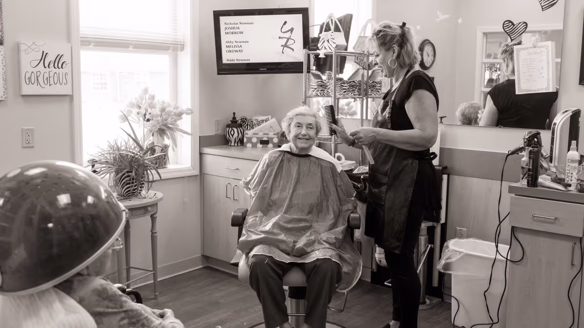 An elderly woman sitting in a salon chair wearing a hairdressing cape, smiling while a hairstylist stands beside her holding a comb and a hairdryer. The room has a window with plants on a small table, a wall-mounted TV, a mirror, and various hair salon tools and products on counters.