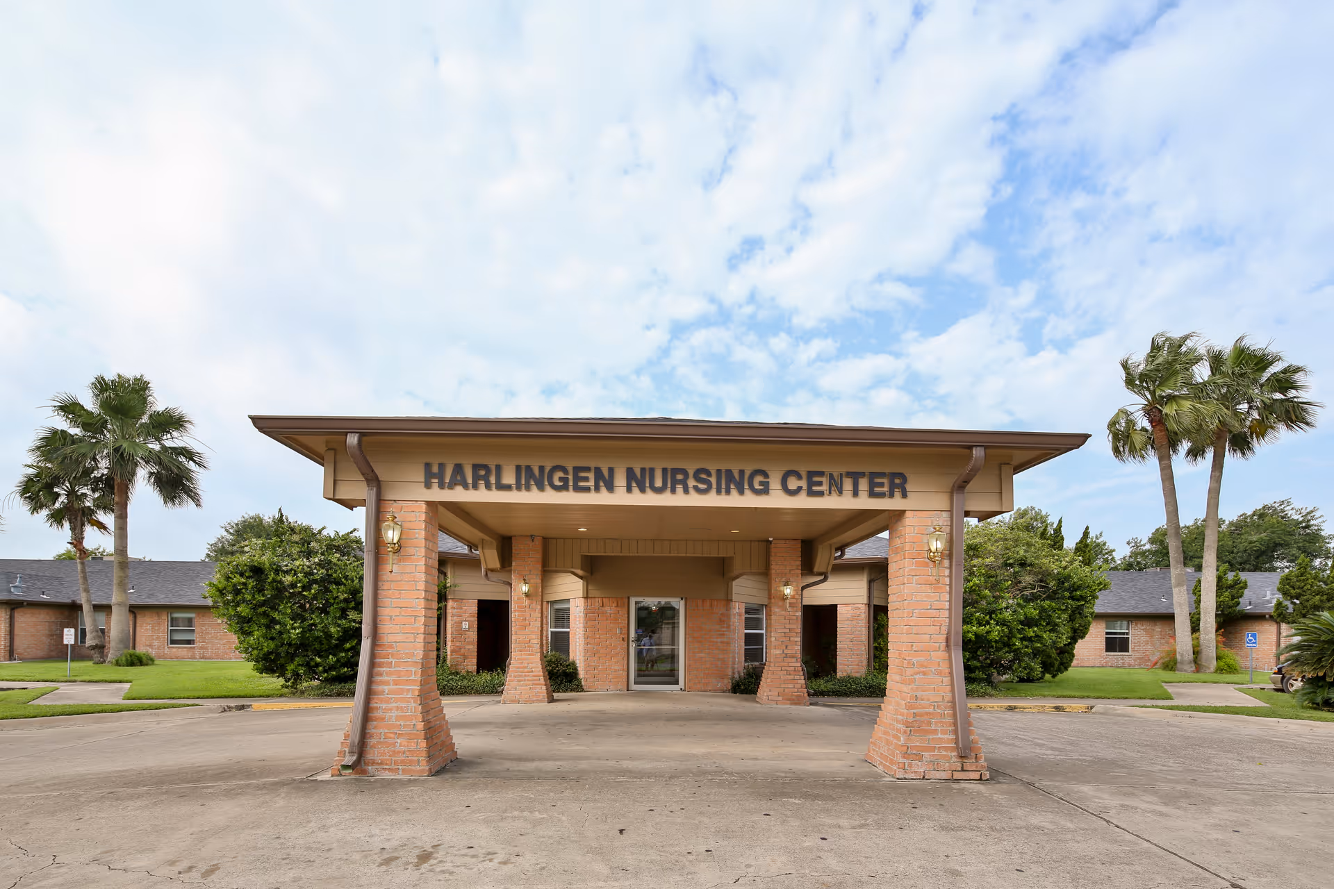 Front exterior view of Harlingen Nursing Center showing a covered entrance supported by brick columns, with palm trees and greenery on either side under a partly cloudy sky.