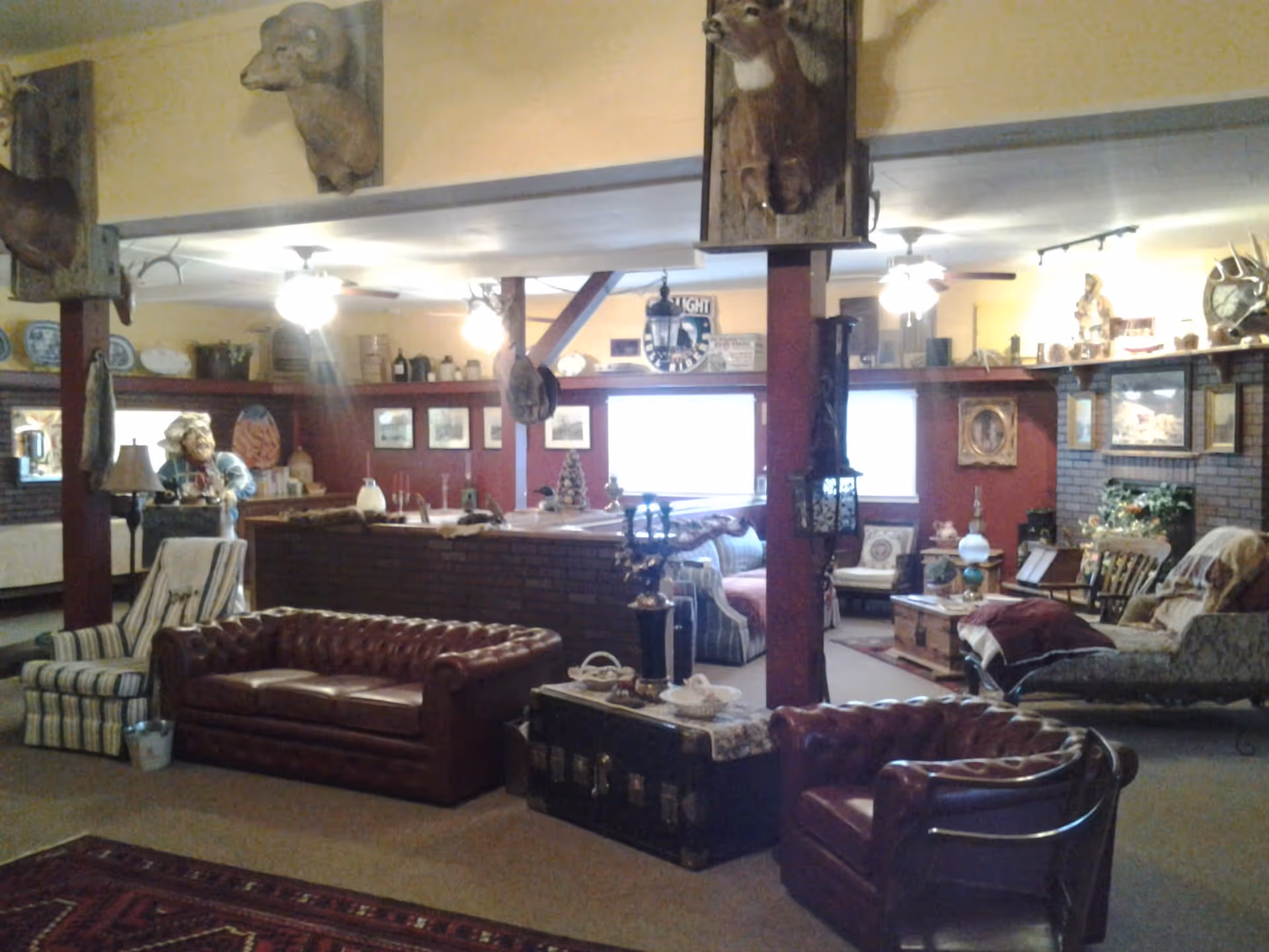 A cozy living room area with vintage and rustic decor, including leather sofas, armchairs, a wooden chest used as a coffee table, mounted animal heads on the walls, and various decorative items on shelves and tables. The room has warm lighting and ceiling fans.