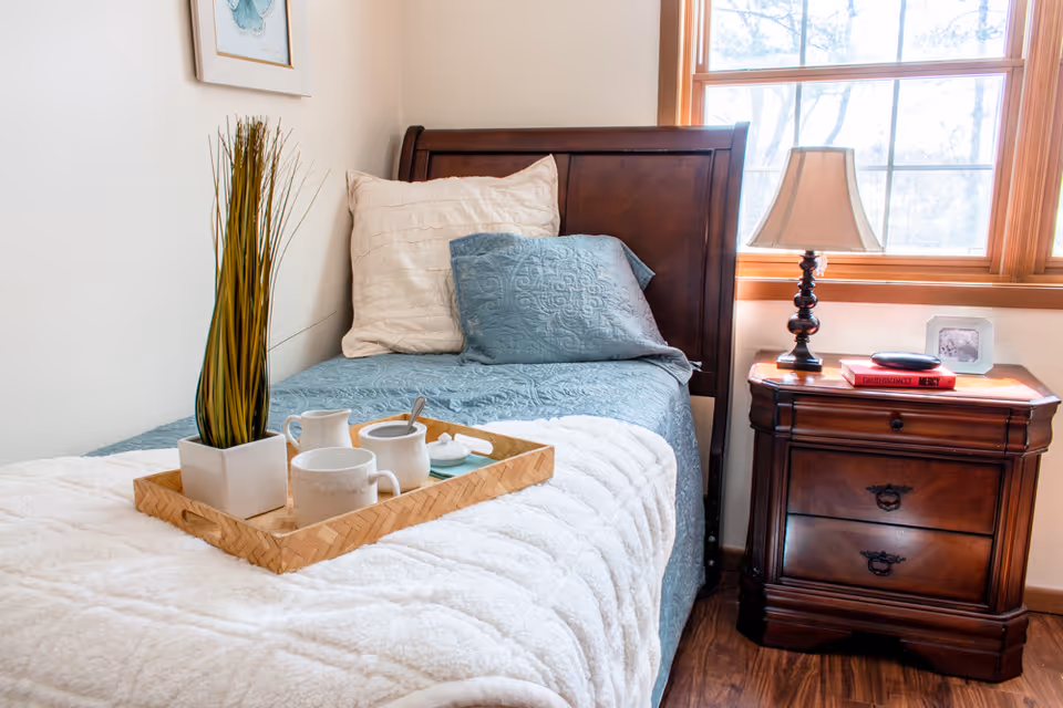 A neatly made single bed with a wooden headboard, decorative pillows, a tray holding cups and a plant, and a wooden bedside table with a lamp by a window.
