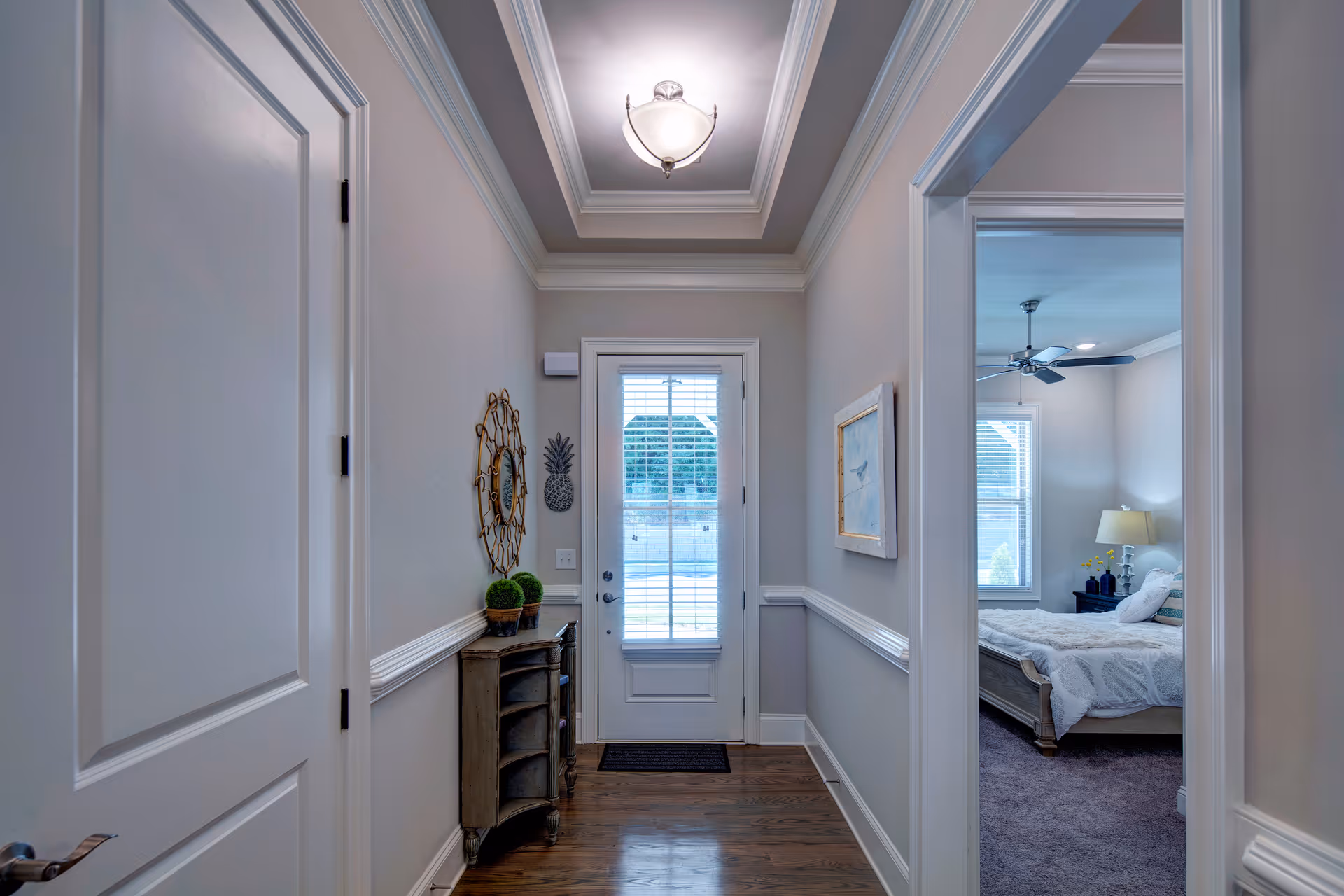 A hallway with wooden flooring and light gray walls featuring white trim. On the left side, there is a closed white door and a small wooden cabinet with decorative plants and a round mirror above it. At the end of the hallway, there is a glass door with blinds allowing natural light to enter. On the right side, an open doorway reveals a bedroom with a bed, nightstand, lamp, ceiling fan, and two windows with blinds.