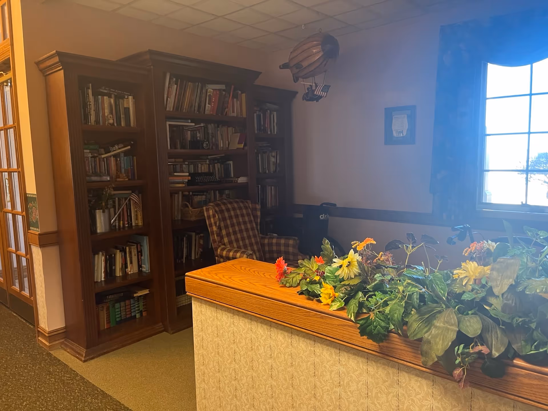 Interior of a cozy room with wooden bookshelves filled with books, a checkered armchair, a window with floral curtains, and a wooden counter adorned with a variety of colorful artificial flowers and green plants.