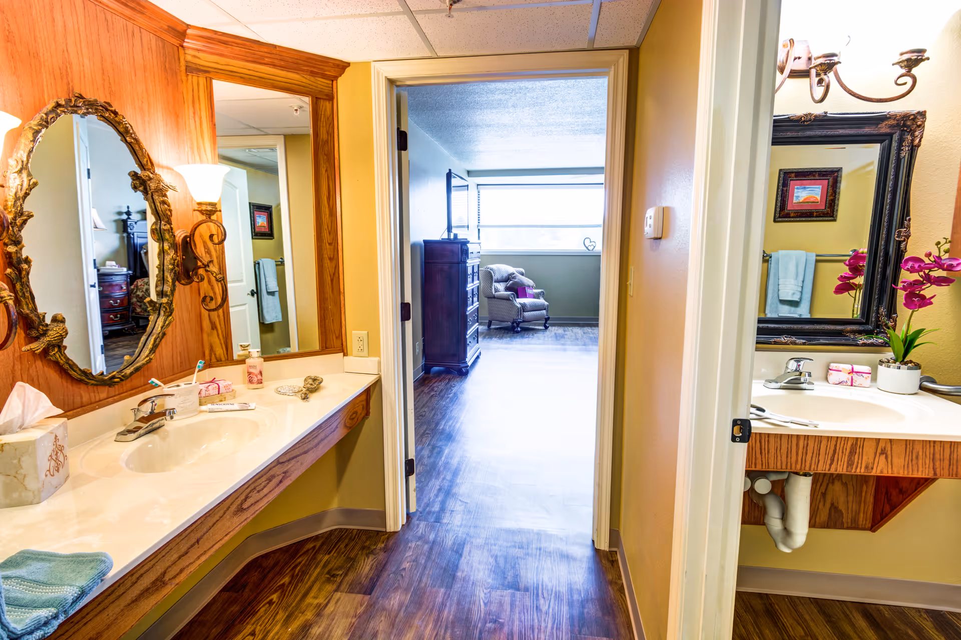 Double-sink bathroom vanity with ornate mirrors and lamps, looking through a doorway into a sitting room with an armchair.
