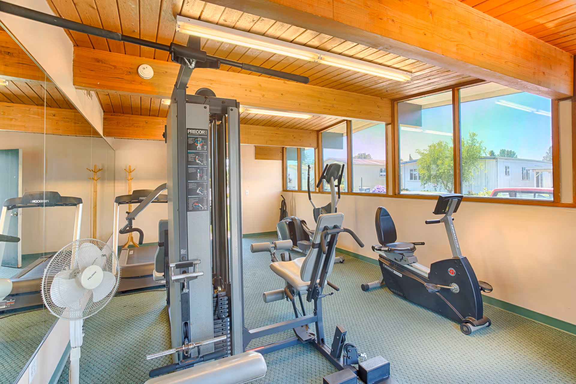 A small fitness room with exercise equipment including a multi-function weight machine, a recumbent stationary bike, and a treadmill. The room has large windows letting in natural light, wooden ceiling beams, and a wall mirror reflecting the equipment. A white standing fan is also visible.