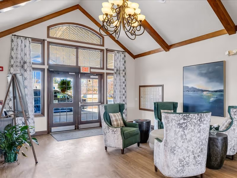A bright and welcoming lobby area with high vaulted ceilings and wooden beams. There are large glass double doors with an exit sign above, flanked by tall windows with patterned curtains. The seating area includes four upholstered chairs in green and gray tones arranged around small round tables. A large abstract painting hangs on the wall, and a set of mailboxes is visible in the background. A chandelier with multiple lights hangs from the ceiling.