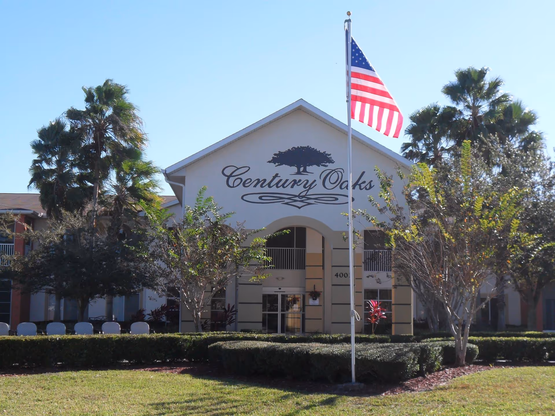 Exterior front view of the Century Oaks senior living facility building with an American flag on a flagpole in the foreground, surrounded by palm trees and other greenery under a clear blue sky.